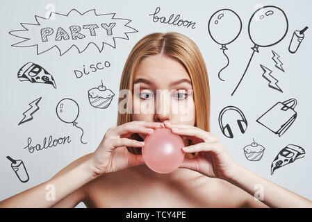 Preparazione per la festa. Giocoso ragazza bionda soffia un palloncino rosa mentre in piedi contro uno sfondo grigio con party tema scarabocchi su di esso. Avendo divertimento. Rullo di trascinamento ausiliario Foto Stock