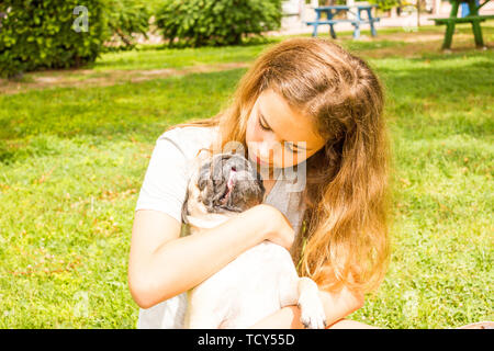 Giovane ragazza adolescente avvolge il suo cane pug nel parco su erba verde Foto Stock