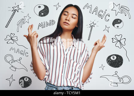 Magic nell'aria. Ritratto di sognante asian giovane donna in abbigliamento casual incrocio le dita mentre in piedi contro uno sfondo grigio con disegnati a mano la magia per Foto Stock