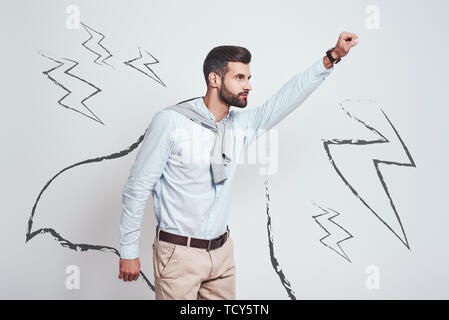 Come un eroe. Giovane uomo barbuto che indossa un capo disegnato e sollevando la sua una mano mentre in piedi contro uno sfondo grigio con illustrazione del lightn Foto Stock