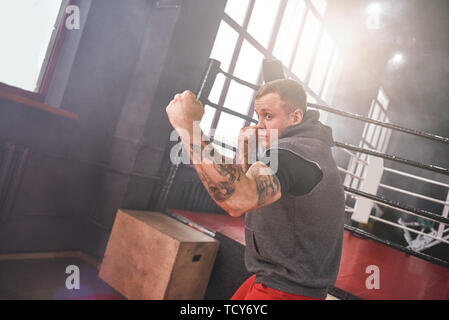 Fiducioso bello atleta in abbigliamento sportivo gettando uppercut. Giovane uomo boxe con ombra mentre in piedi di fronte colorata anello di inscatolamento Foto Stock