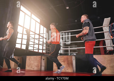 Un gruppo di smilling muscolare focalizzato sportmen in fase di preriscaldamento e in esecuzione nei pressi di pugilato ring prima dell'allenamento in palestra Foto Stock