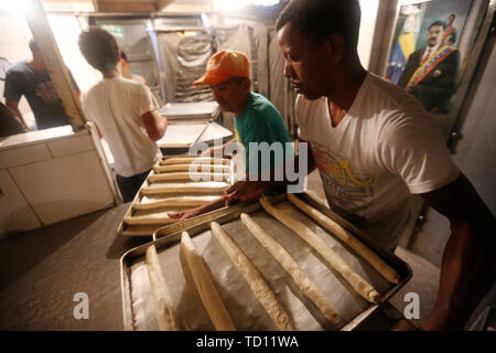 05 giugno 2019, Venezuela, Caracas: i dipendenti portano vassoi da forno con baguette in 'La Minka', un panificio gestito da Maduro sostenitori del governo. Sullo sfondo si può vedere un poster con una foto del presidente. L'ex panificio privato è stato chiamato 'Mansion's Bakery'. Ora circa 6.000 pezzi di pane sono prodotte ogni giorno a prezzi fissati dallo stato. Il paese con la più grande riserva di petrolio nel mondo è in un drammatico declino. La svalutazione del denaro è il più alto del mondo, per molte persone il cibo è insostenibile. Questi "' Bolivariana panetterie, che vendono il pane a prezzi fissi, vedere loro ta Foto Stock