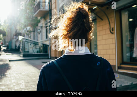 Vista posteriore del giovane donna camminare per la strada, sun flare verso di lei. Femmina con ricci di acconciatura di coda di cavallo passa lungo il marciapiede della città alla giornata di sole. Foto Stock