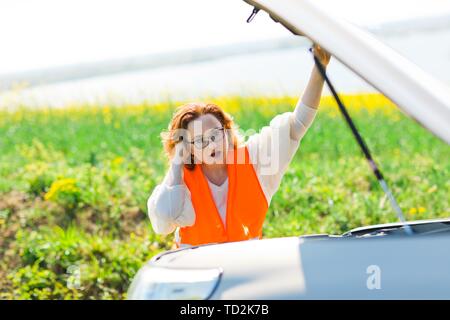 Una donna in arancione vest auto aperto il cofano della vettura rotto - cercando di risolvere il problema Foto Stock