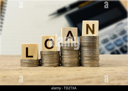 Prestito finanziario concetto. Blocco di legno sulle monete impilate in crescita su tavola di legno con notebook sfocate, penna e calcolatrice. Mostra prestare e prendere in prestito per pagare Foto Stock
