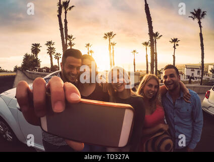 Happy amici in piedi nella parte anteriore della vettura convertibile e tenendo selfie con la fotocamera del cellulare - il giovane popolo millenario divertirsi su roadtrip Foto Stock