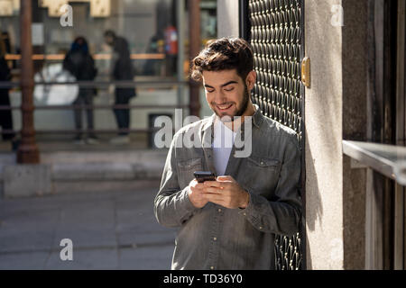 Moda uomo college nel suo ventenne felice controllo blog o chattare su internet con amici su smart telefono mobile app in unione all'aperto della città. Foto Stock
