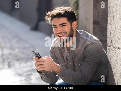 Moda uomo college nel suo ventenne felice controllo blog o chattare su internet con amici su smart telefono mobile app in unione all'aperto della città. Foto Stock