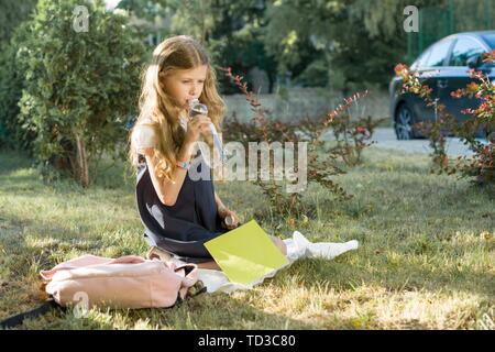 Ragazza in uniforme scolastica con zaino seduto sul prato in cantiere e acqua potabile da bottiglia nelle calde giornate di sole Foto Stock