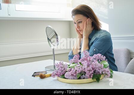 Ritratto di donna matura con specchio per il make-up il massaggio viso e collo, bella femmina 40 anni seduto a casa a tavola con fiori lilla Foto Stock