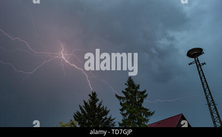 Sieversdorf, Germania. 12 Giugno, 2019. Molti fulmini di un temporale illumina il cielo di sera al di sopra di una cicogna il nido e una casa del tetto. Credito: Patrick Pleul/dpa-Zentralbild/dpa/Alamy Live News Foto Stock