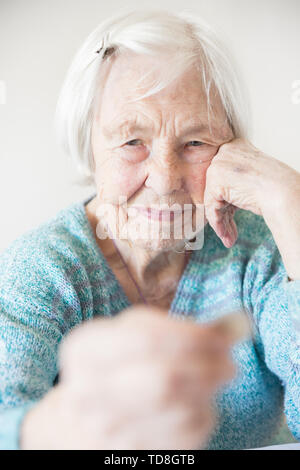 Triste anziani donna seduta al tavolo a casa e guardando miseramente solo al restante moneta dalla pensione nella sua mano. Foto Stock