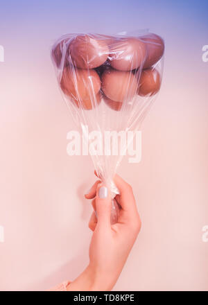 Le uova in un sacchetto di plastica capovolto come palloncini nella donna la mano sul multi colore blu e corallo chiaro sfondo rosa con luce morbida Foto Stock
