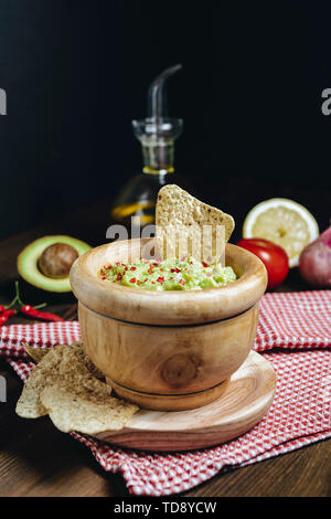 Guacamole deliziosi in una ciotola di legno con tortilla chips accanto a avocado e altri ingredienti, messicana tipica sana cucina vegana con rustici dark f Foto Stock