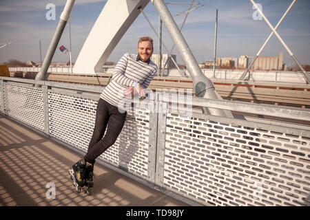 Un giovane uomo, ponendo in piedi a una telecamera, sorridente cercando felice indossando pattini inline. all'aperto su un ponte è un giorno di sole. Foto Stock