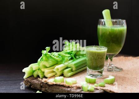 Mescolate il succo di sedano in drink di benvenuto il vetro e il mazzetto di sedano fresco levetta sul tavolo in legno con foglie su sfondo nero. Alimenti e ingredienti di Foto Stock