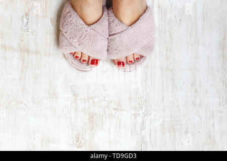 Gambe femmina rosso con chiodi in home pellicce soffici pantofole rosa su una luce sullo sfondo di legno. flat laici. Vista dall'alto. Il concetto di un accogliente luminosa casa ragazza. Foto Stock