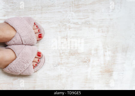 Gambe femmina rosso con chiodi in home pellicce soffici pantofole rosa su una luce sullo sfondo di legno. flat laici. Vista dall'alto. Il concetto di un accogliente luminosa casa ragazza. Foto Stock