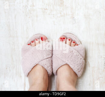 Gambe femmina rosso con chiodi in home pellicce soffici pantofole rosa su una luce sullo sfondo di legno. flat laici. Vista dall'alto. Il concetto di un accogliente luminosa casa ragazza. Foto Stock