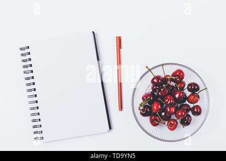 Vista superiore della scrivania con notebook vuota e penna accanto a snack sani di ciliegie fresche, piatto giaceva su sfondo bianco. Foto Stock