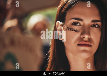 Primo piano di una giovane attivista con la parola libertà scritta sul viso guardando la macchina fotografica mentre protestava all'aperto con un gruppo di dimostranti. Foto Stock
