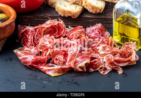 Stuzzicanti fette di prosciutto iberico in primo piano. Olio d'oliva, il pane e il pomodoro fresco, olive. Sfondo nero. Rustico e aspetto artigianale. Foto Stock