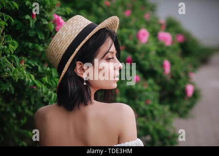 Bella ragazza in estate hat guardando rose fiori con sorriso. Modello in posa vicino a fioritura di rose. Foto Stock