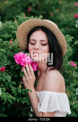 Romantico close-up verticale o affascinante bruna ragazza nel cappello di paglia odori fiori sul giardino delle rose Foto Stock