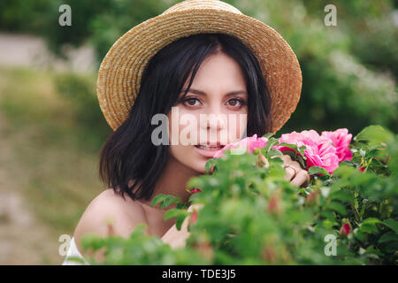 Romantico close-up verticale o affascinante bruna ragazza nel cappello di paglia odori fiori sul giardino delle rose Foto Stock