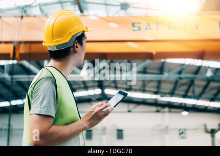 Giovani asiatici pesante maschio industral lavoratore utilizzando una tavoletta digitale all'interno della fabbricazione e del sito di fabbrica con gru dietro Foto Stock