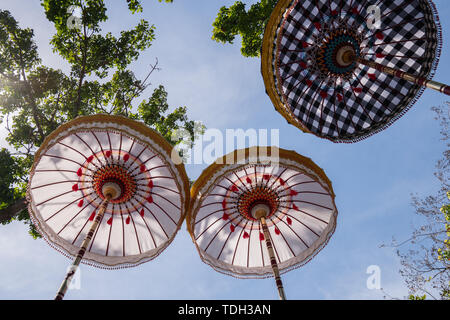 Gruppo di coloratissimi ombrelli Balinese alla cerimonia di celebrazione nel tempio indù. Design tradizionale, bianco e motivo a scacchi sotto l'albero Foto Stock