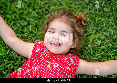 Sorridenti poco Toddler Girl posa su erba nel parco con abito rosso. Foto Stock