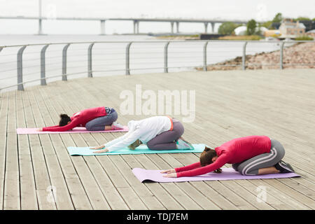 Vista laterale ritratto di tre donne contemporanee a praticare lo yoga all'aperto e stretching all'alba dal fiume, spazio di copia Foto Stock