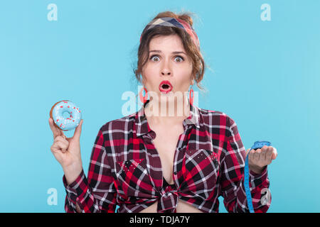 La dieta, la perdita di peso e il cibo spazzatura - pin-up donna scegliendo tra il dessert e il nastro di misurazione su sfondo blu Foto Stock