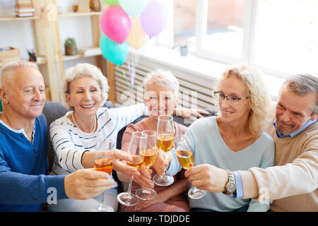 Il gruppo di allegro coppia amici entusiasti di festa di compleanno seduto sul divano nel soggiorno e flauti di sollevamento Foto Stock