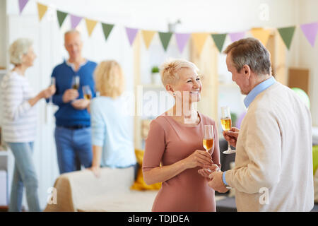Positivo attraente donna matura con capelli corti in piedi nel soggiorno e a parlare con l'uomo in house party, essi bevendo champagne e flirtare Foto Stock