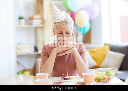 Grave pensieroso donna anziana in party hat seduta a tavola e guardare la torta di compleanno mentre festeggia il compleanno da solo Foto Stock