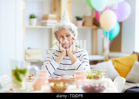 Grave pensionato infelice grey-haired lady in party hat seduti al tavolo da pranzo pieno di piatti e guardando alla torta di compleanno con candela pur avendo bir Foto Stock