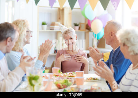 Gruppo positivo della coppia amici seduti al tavolo da pranzo in soggiorno e battendo le mani per il compleanno della donna mentre congratularmi con lei insieme Foto Stock