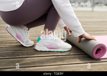 Primo piano della donna contemporanea bianco da indossare calzature sportive rolling materassino yoga sul molo in legno all'aperto, spazio di copia Foto Stock