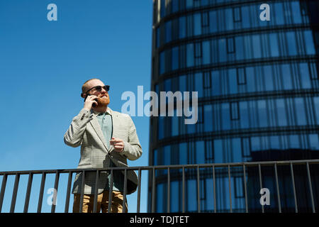 Positivo bello mirato barbuto manager in occhiali da sole in piedi contro il grattacielo e avente la conversazione telefonica Foto Stock