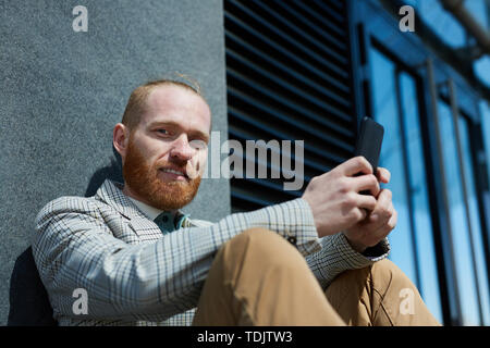 Ritratto di sorridere bello barbuto imprenditore della camicia casual seduti sul suolo e utilizzando il telefono Foto Stock