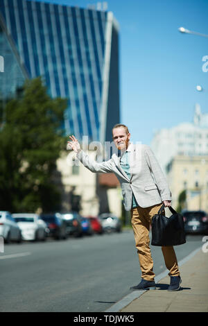 Positivo bel giovane imprenditore con valigetta agitando la mano mentre la cattura di taxi sulla strada Foto Stock