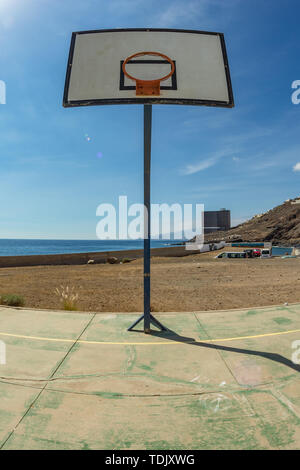 La pallacanestro tavola spinale con cestello sul vecchio campo sportivo. Un enorme edificio abbandonato di fronte all'oceano nel lontano sullo sfondo. Tenerife. Grandangolo, f Foto Stock