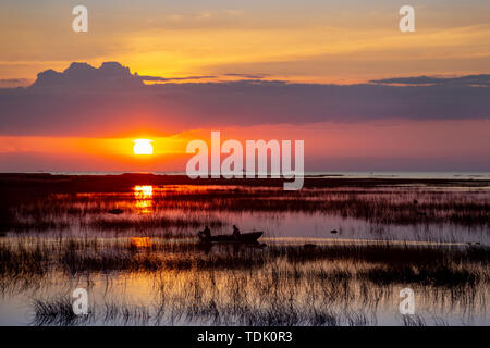 Silhouette di una barca da pesca con due pescatori, galleggiante a remi sulle calme e limpide acque del lago, coperto con sedge al tramonto sullo sfondo di un bel tramonto multicolore cielo nuvoloso riflessa nell'acqua. Foto Stock