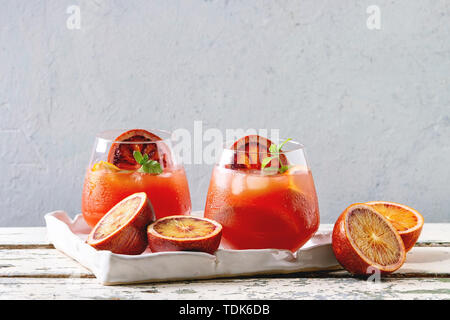 Arancio sanguigno cocktail ghiacciato in bicchieri, decorata da fetta di arance e menta fresca, servita nel vassoio in ceramica bianca sul tavolo di legno con parete grigia a b Foto Stock