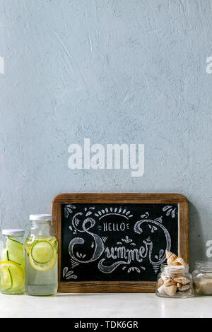 Tema estivo. Chalk lettering Ciao estate sulla lavagna vintage, conchiglie e sabbia in vasetti di vetro, occhiali da sole e due bottiglie di vetro con acqua sassy su Foto Stock