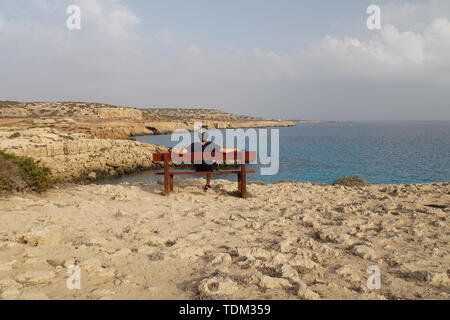 Uomo seduto su un banco di lavoro con vista sul mare. Un uomo si ammira lo splendido paesaggio. Guarda lontano all'orizzonte. Il ragazzo è appoggiato sul banco. Foto Stock