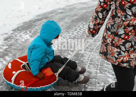 Little Boy su una slitta in inverno. mamma tira il bambino sulla tubazione verso il basso la strada Foto Stock